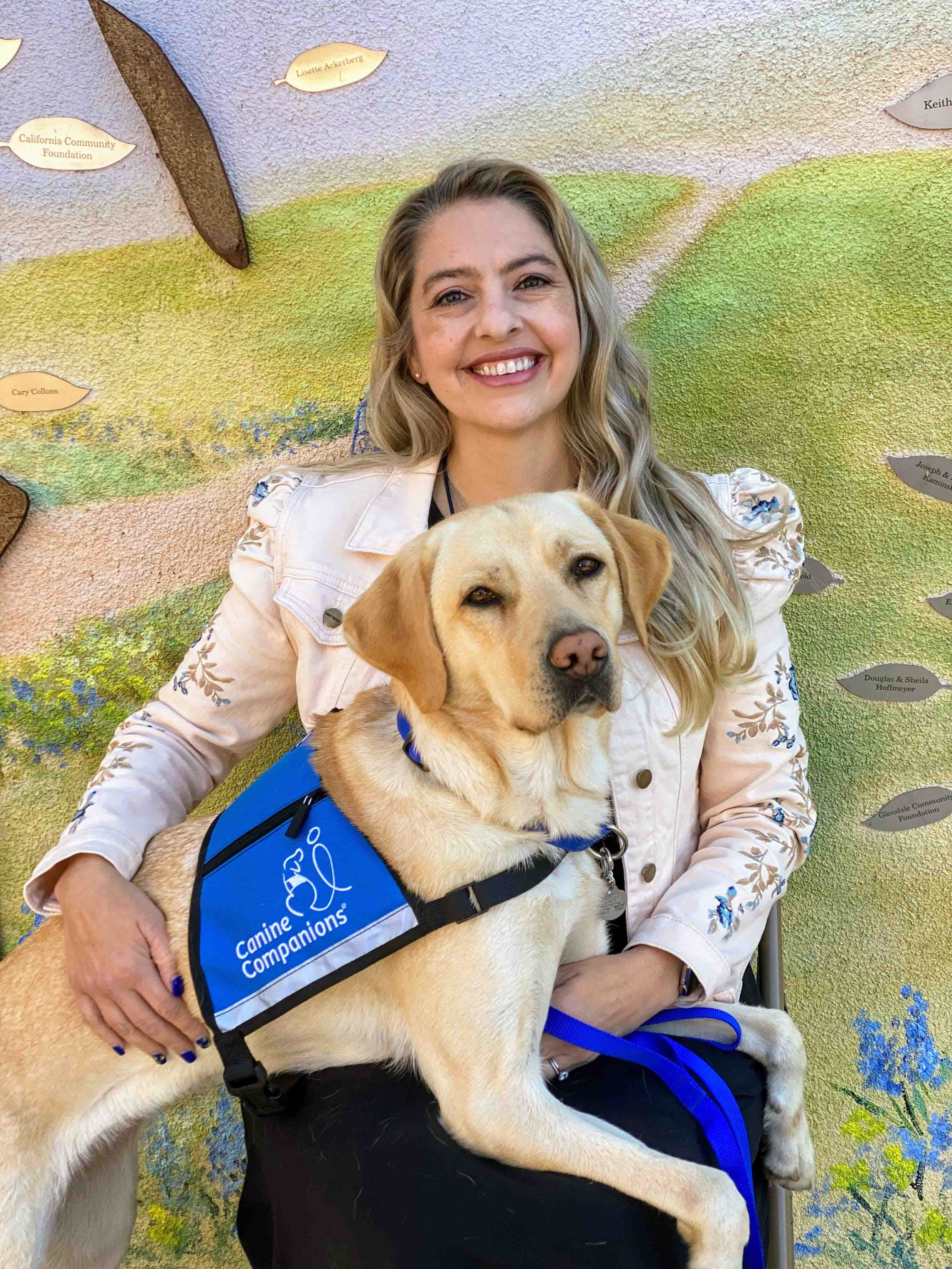 a white woman sits with a yellow lab therapy dog in a vest on her lab