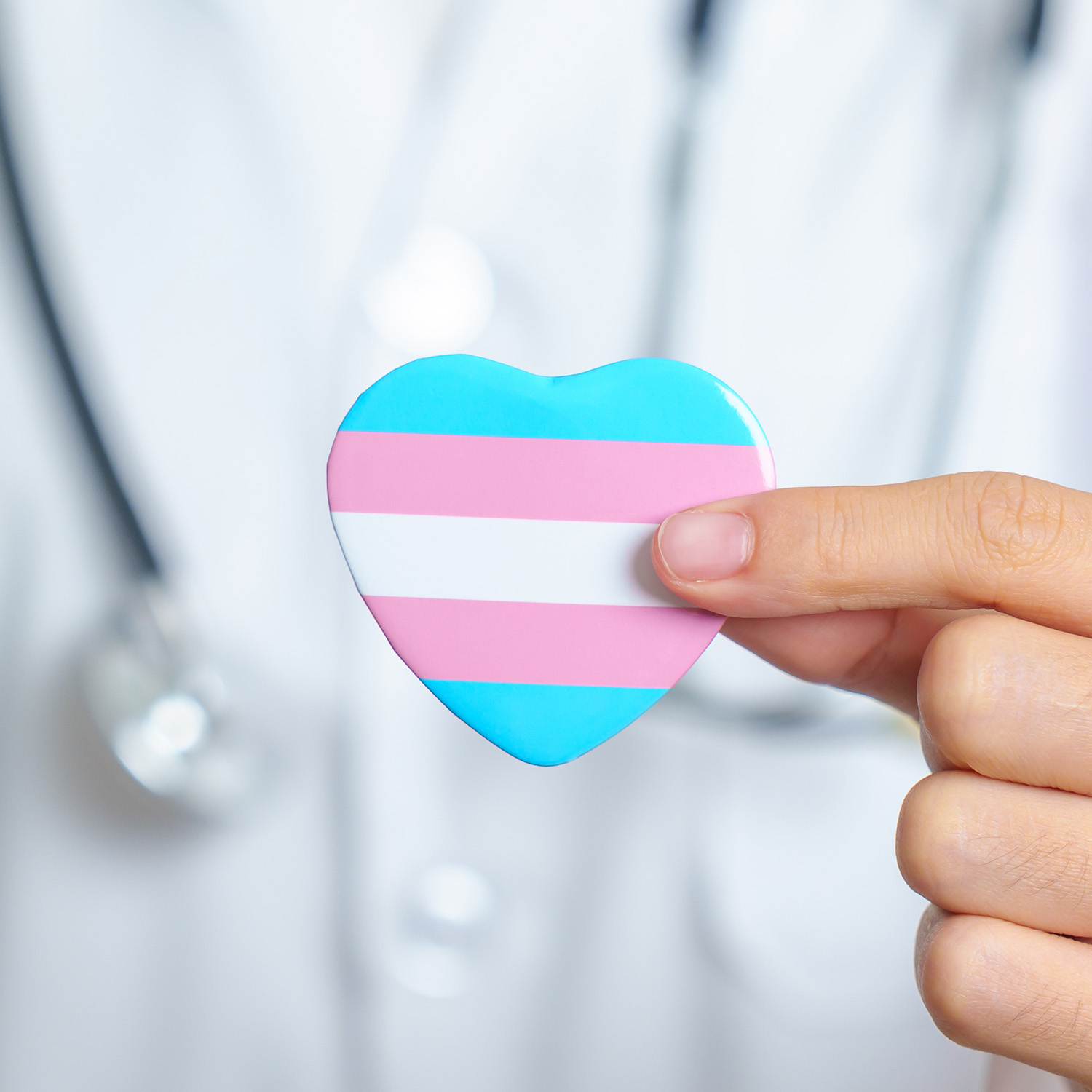 A blue, white, and pink heart being held by a person's hand with a white coat and a stethoscope in the background