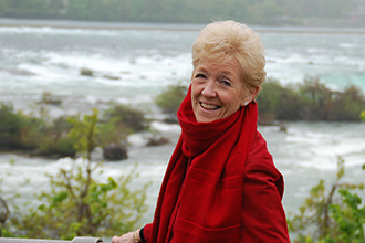 Photo of Judy Watts Brady wearing red and smiling at the camera, with water and shrubs in the background