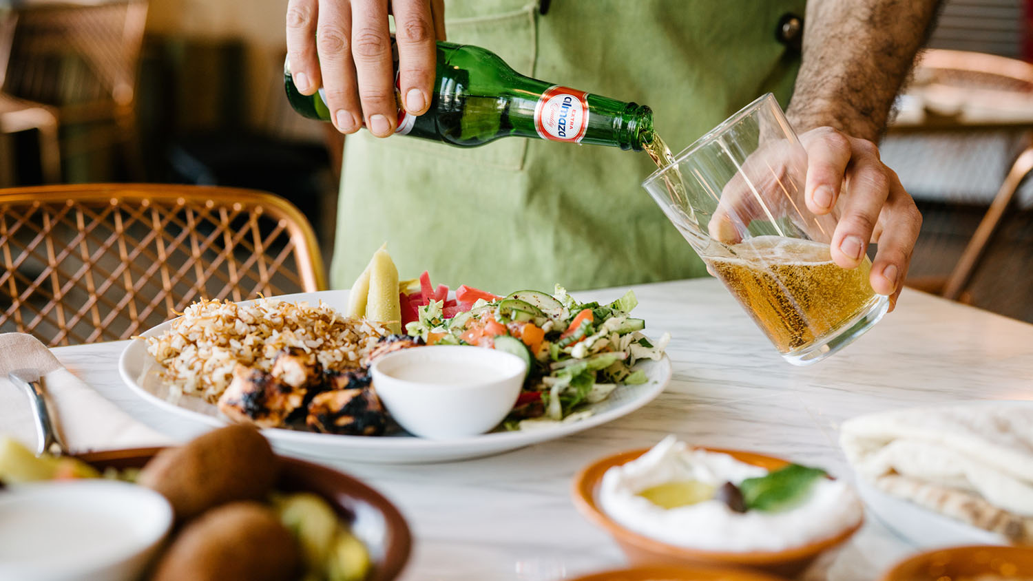 Multiple dishes filled with beautiful Middle Eastern foods in the foreground, with a hand pouring a beer into a glass in the background
