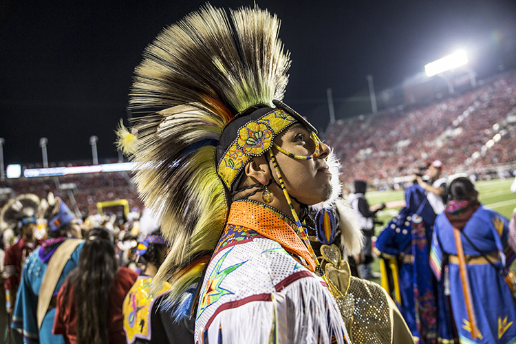 Photo of a Ute performer in traditional dress, standing on the field of Rice Eccles Stadium, surrounded by other Ute dancers