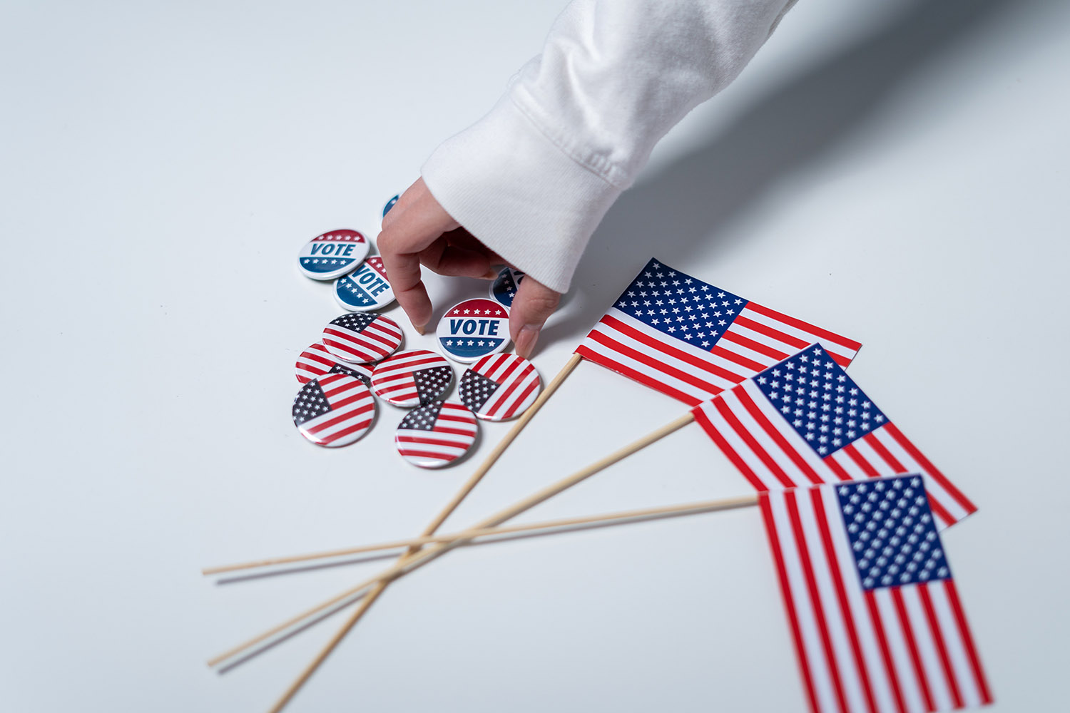 A hand reaches down for a "vote" button, which is sitting on a table with other buttons that say "vote" or have the American flag. Next to the buttons there are three small American flags on wood dowels.