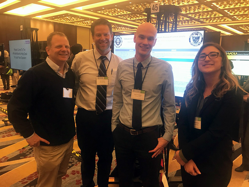 Photo of Dr. Brad Lundahl, Dr. Rob Butters, PhD student Brian Droubay, and MSW student Kalani Nelford in front of an electronic poster