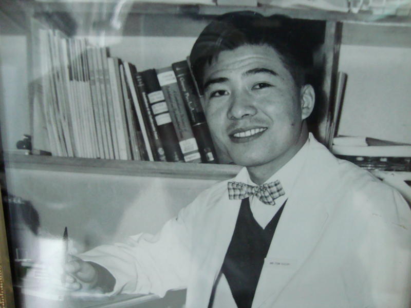 Young Tom Suzuki in a white lab coat, smiling at the camera