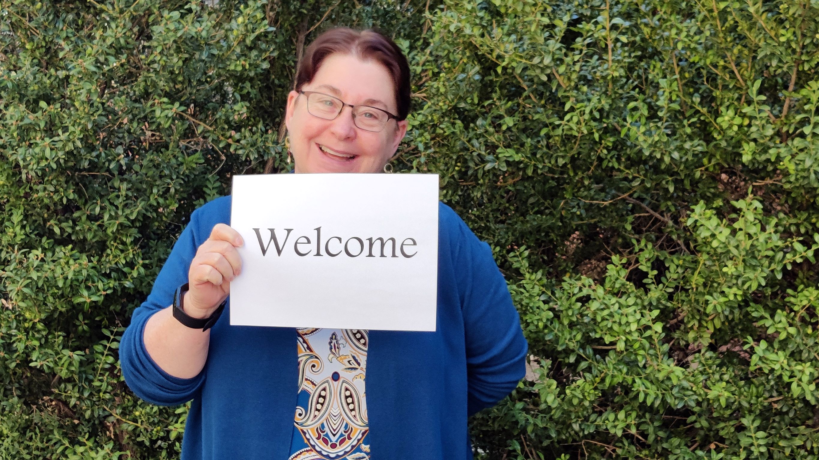 A middle aged white woman wearing glasses is smiling and holding a sign that says "Welcome"