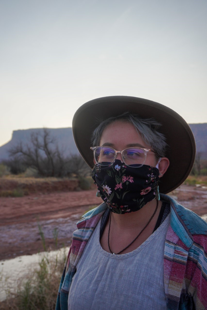 A woman in a hat, wearing a mask, looks off into the distance in a desert landscape