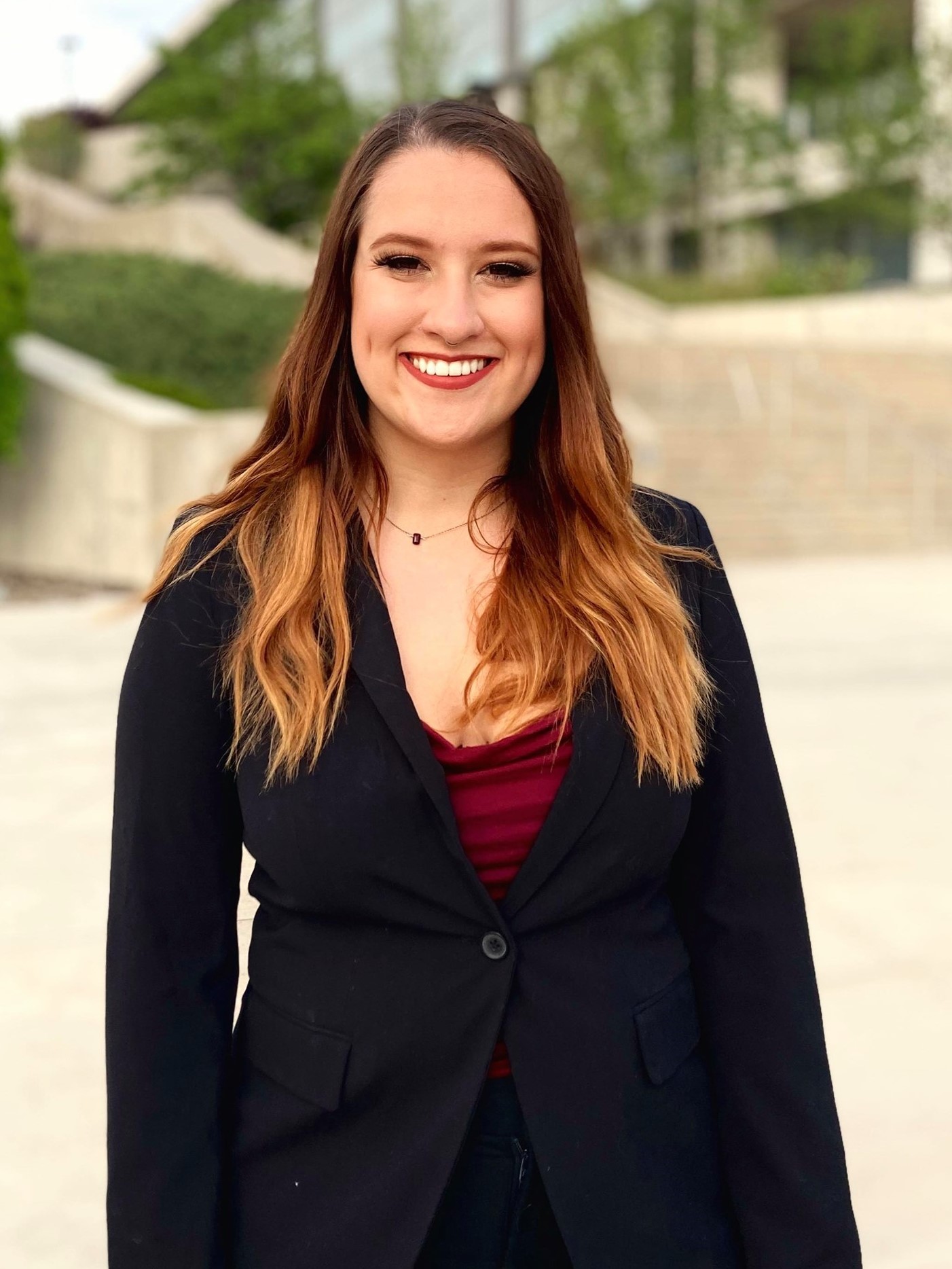 A headshot of a smiling young woman in a black blazer standing outside.