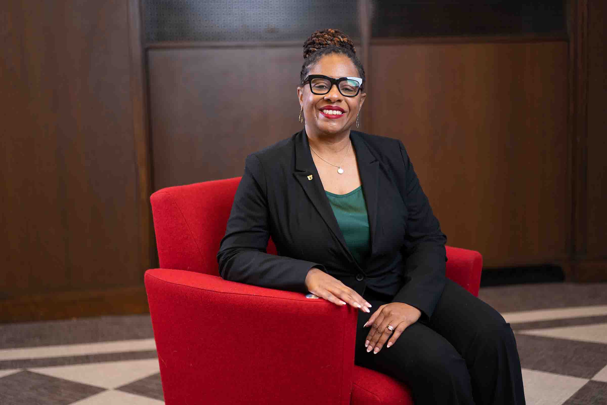 A photo of a middle aged Black woman sitting in a chair, smiling at the camera.