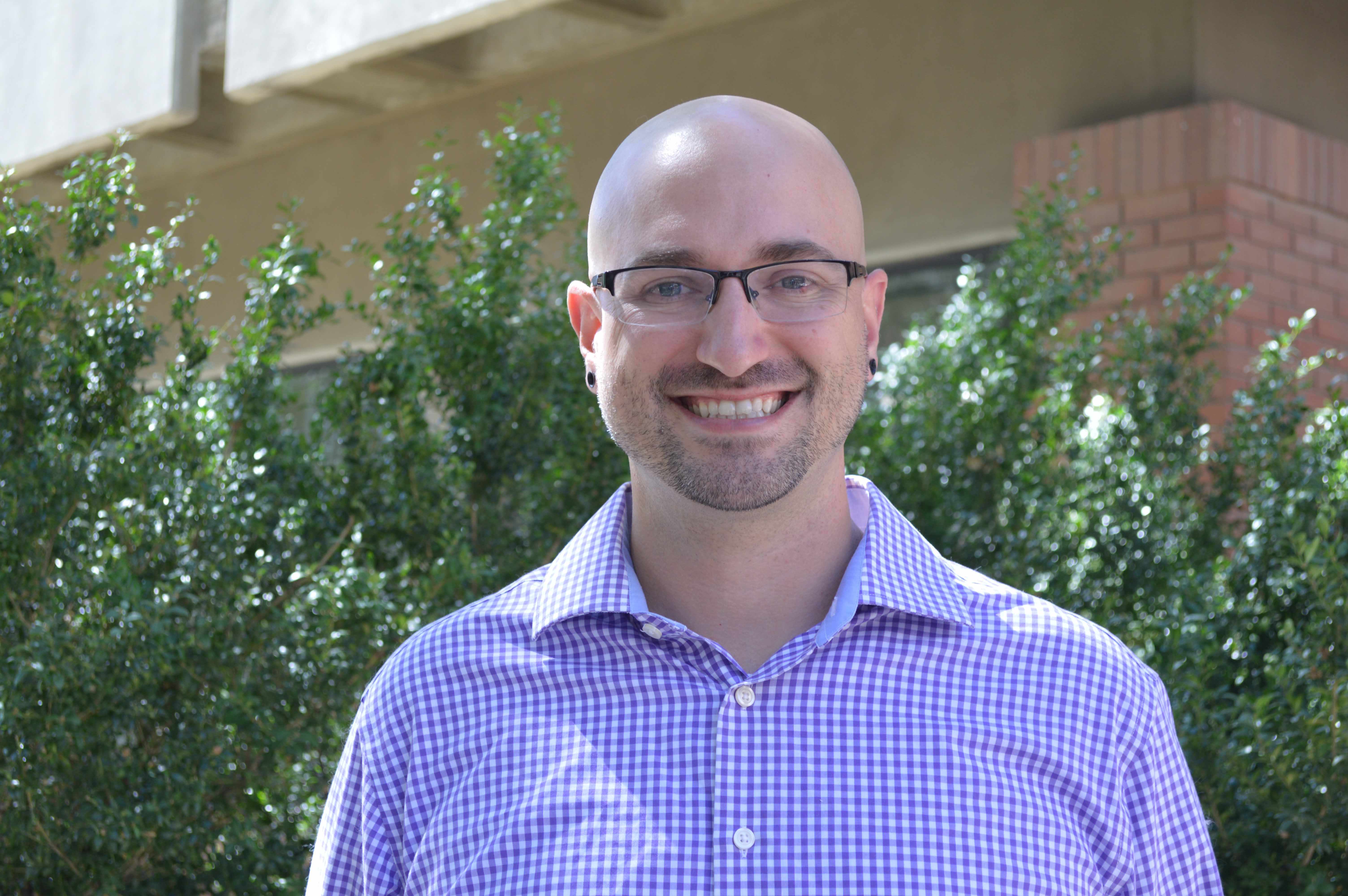 A headshot of a middle aged white man smiling in front of a trees and a brick building.