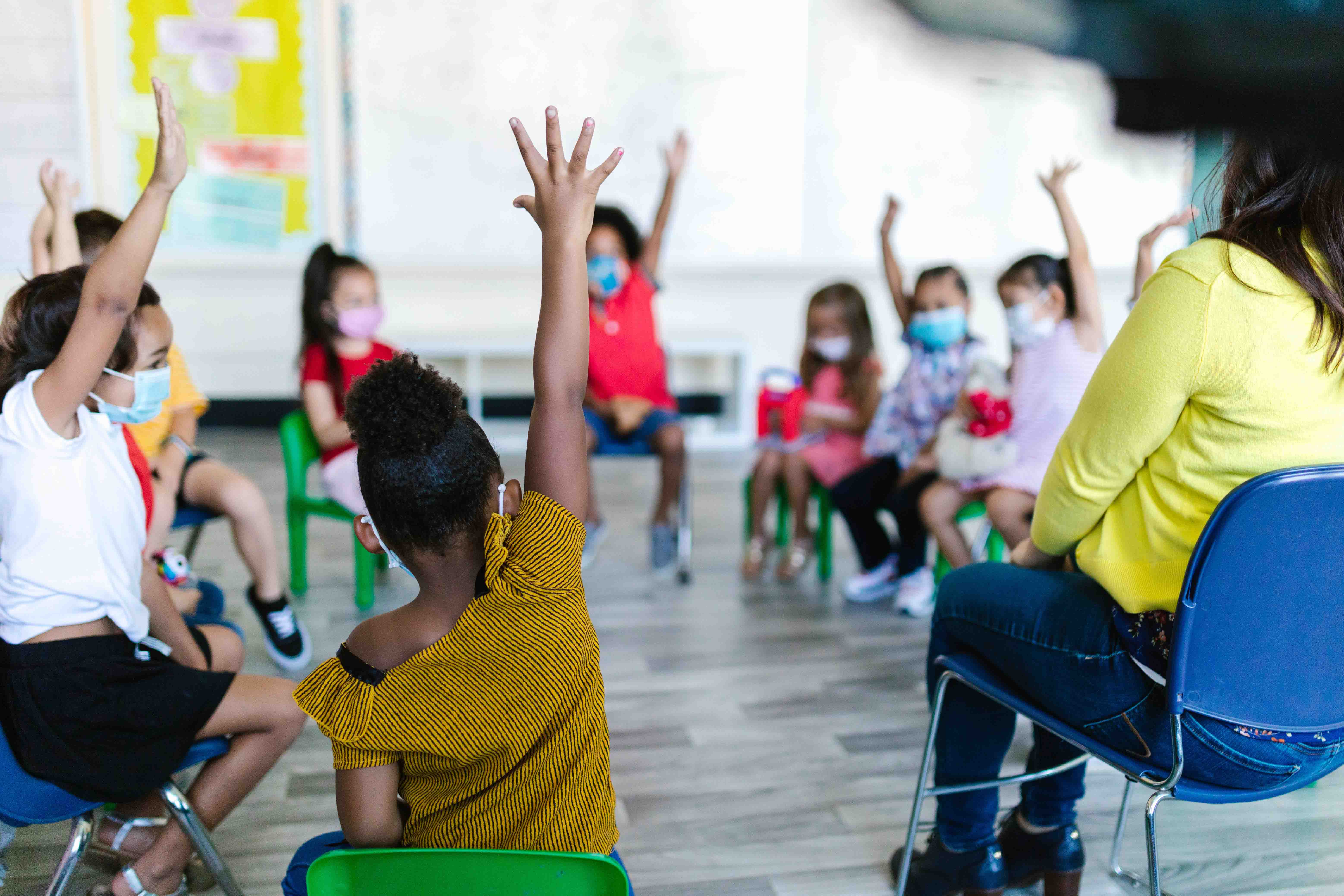 A group of preschool aged children sit in a circle and raise their hands