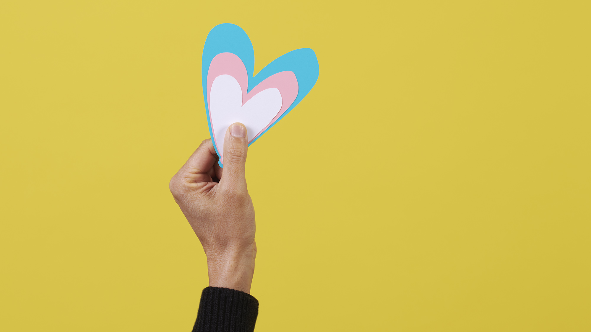 A hand holds up a blue, white, and pink heart in front of a bright yellow background