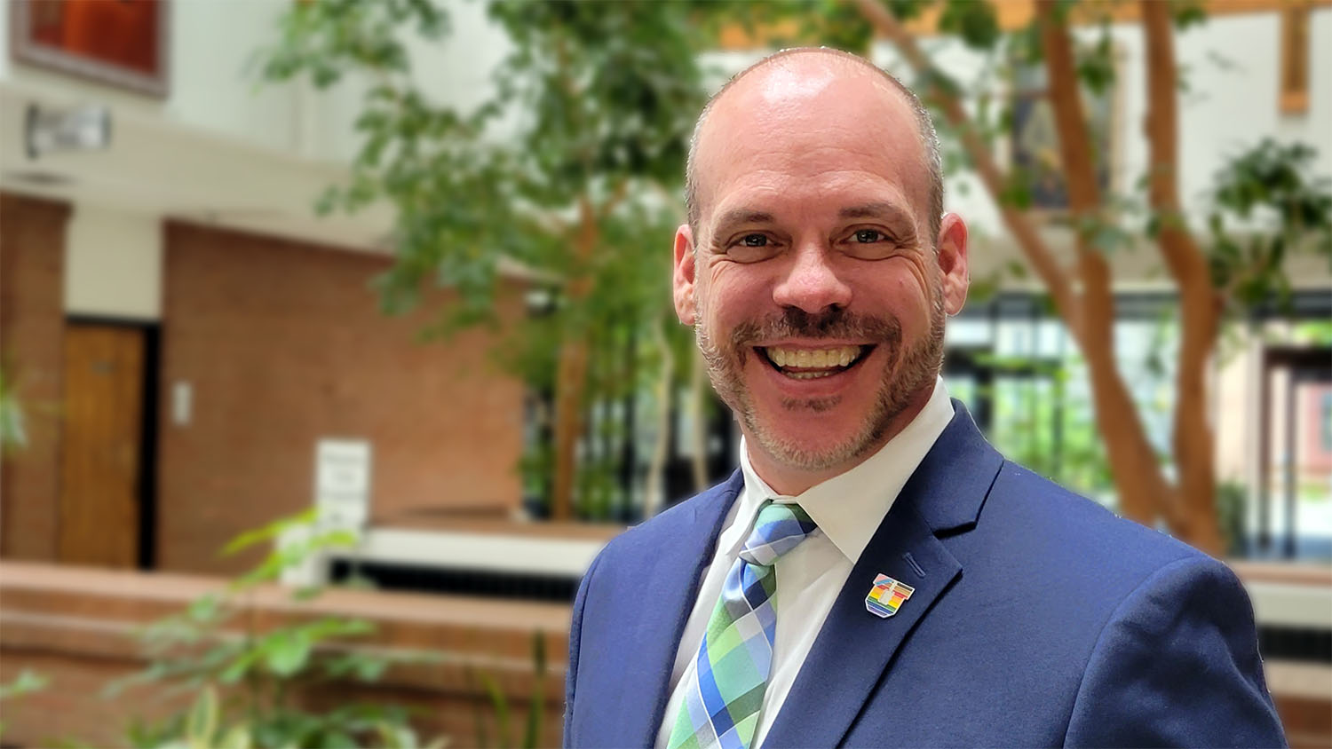 Photo of Dean Philip Osteen, wearing a rainbow U pin and standing in the atrium of the College of Social Work, exuding joy