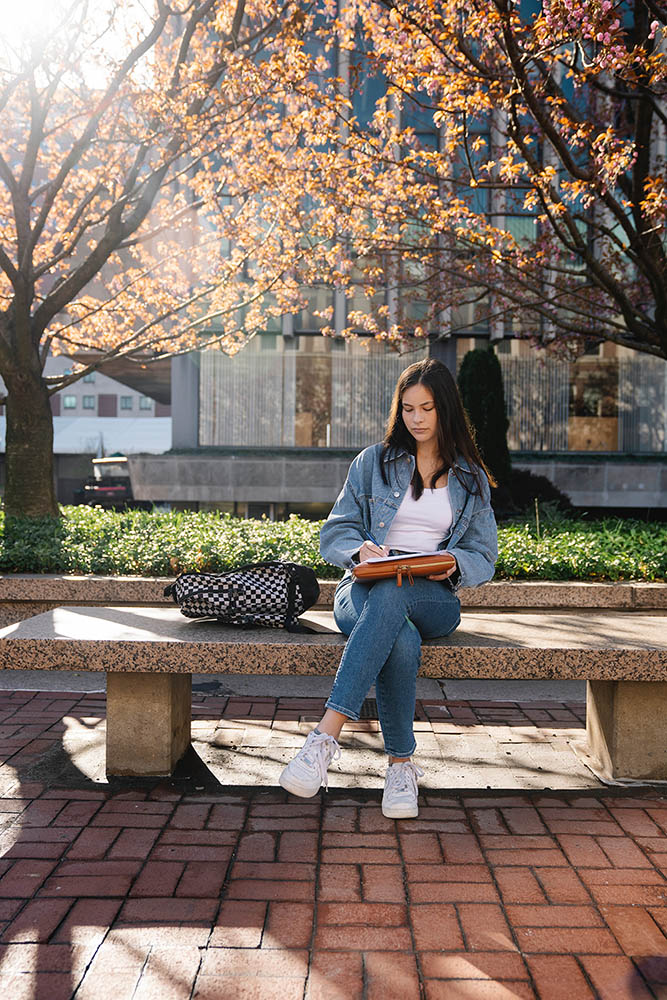 A female student sits on an outdoor bench, writing on a pad, a checkered backpack next to her