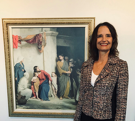 A white woman wearing a blazer smiles at the camera.  She stands in front of a framed photo of Jesus Christ among the poor.
