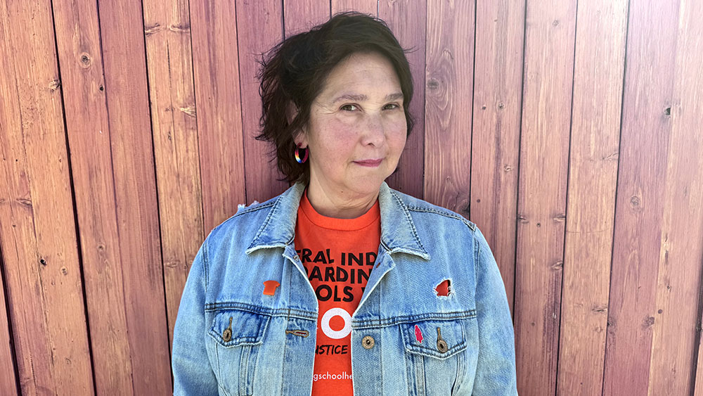 A woman wearing an orange shirt and jean jacket up against a wooden panel background.