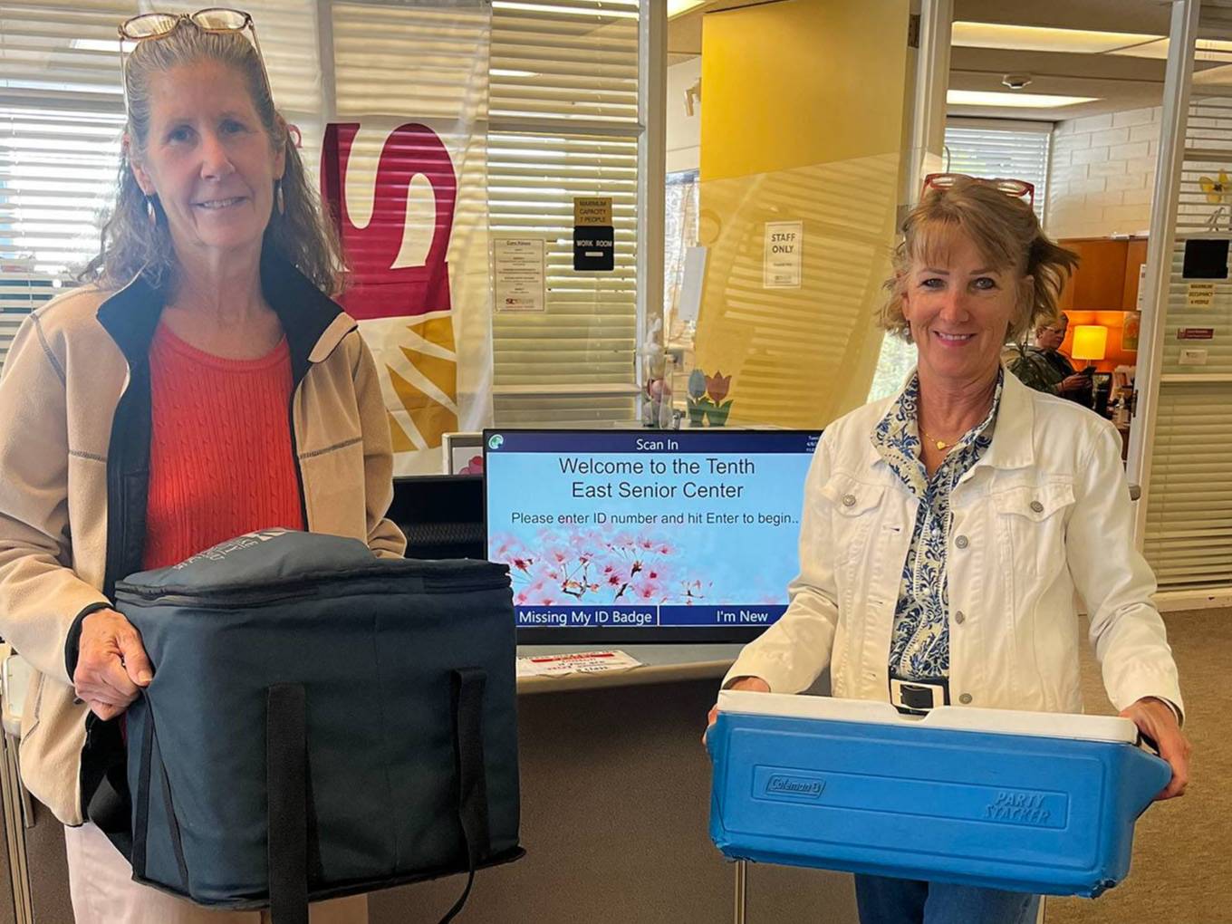Two smiling women standing and holding a bag and a cooler.