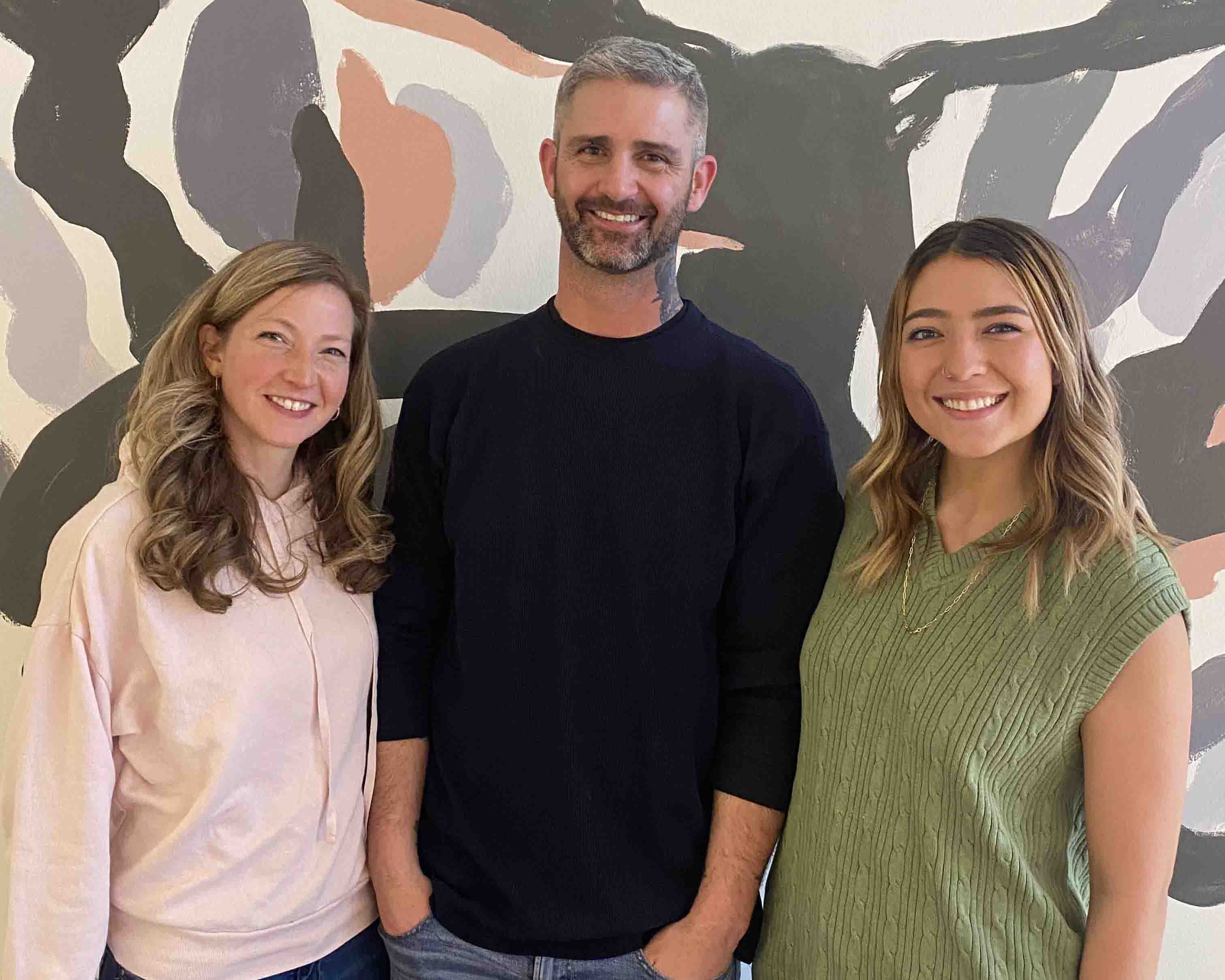 three white people of varying ages smile a the camera in front of a patterned background