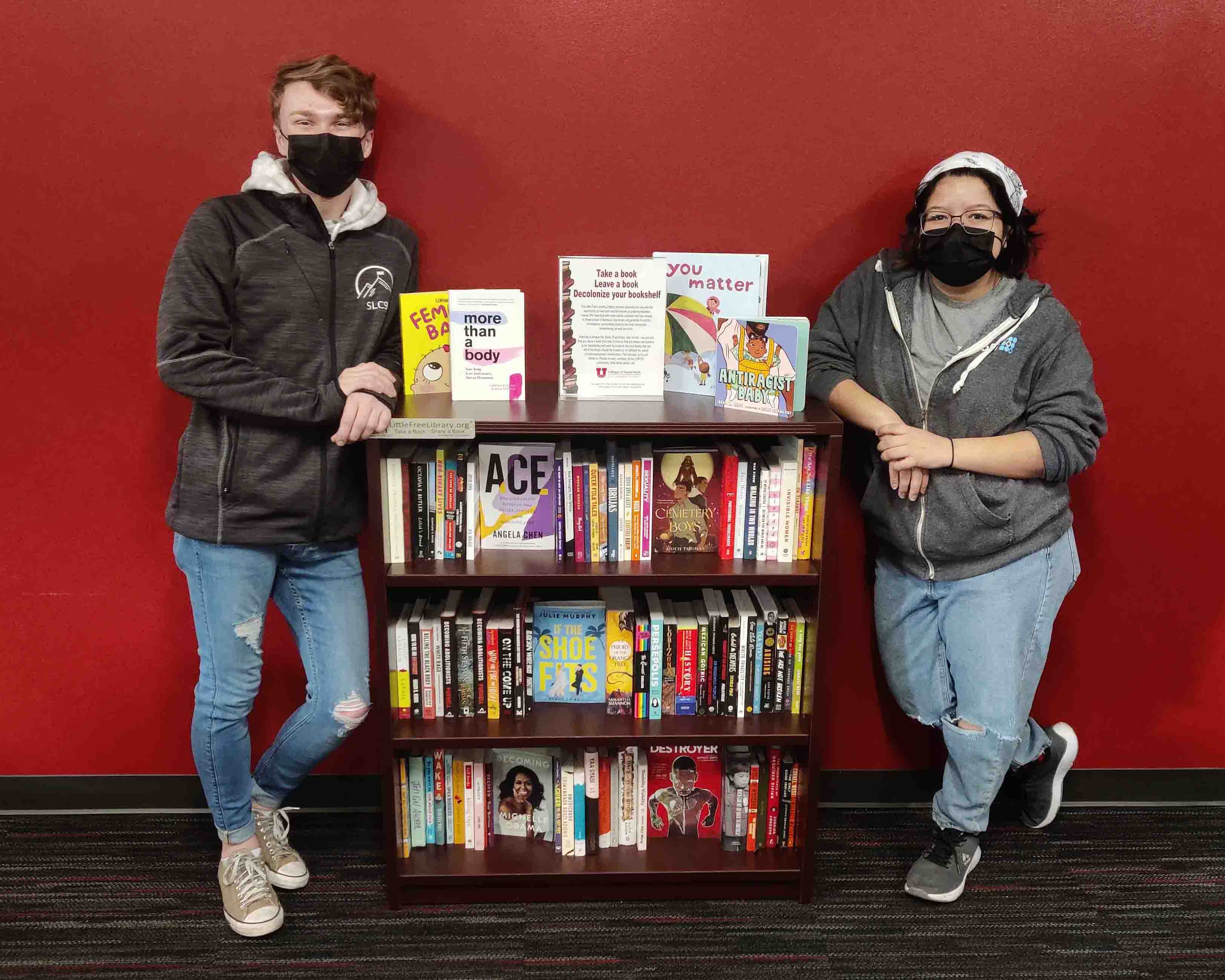 Two masked college students stand on opposite ends of a bookshelf full of books