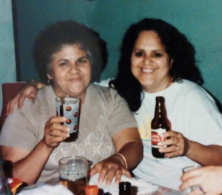 An photo of two Latinx women sitting at a table, smiling at the camera, with drinks in hand.