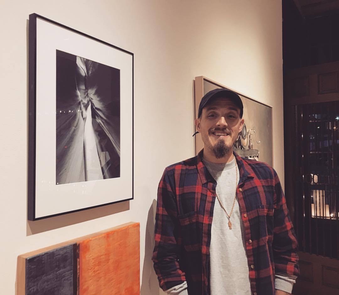 a young white man in a baseball cap and flannel shirt is smiling at the camera while standing in front of an art gallery wall