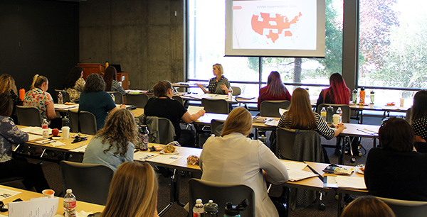 Photo of a woman presenting to a room of seated professionals. A map with certain states highlighted is on a screen in the background.